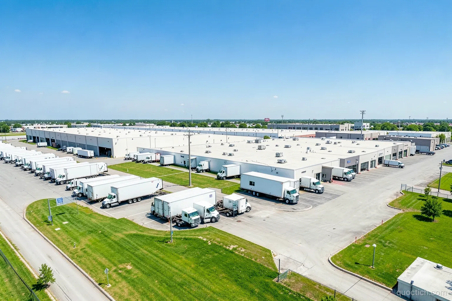 Aerial wide view of a modern logistics warehouse complex with rows of white trucks and green lawns outside Dallas, Texas, bright midday sun, clear blue sky, no people